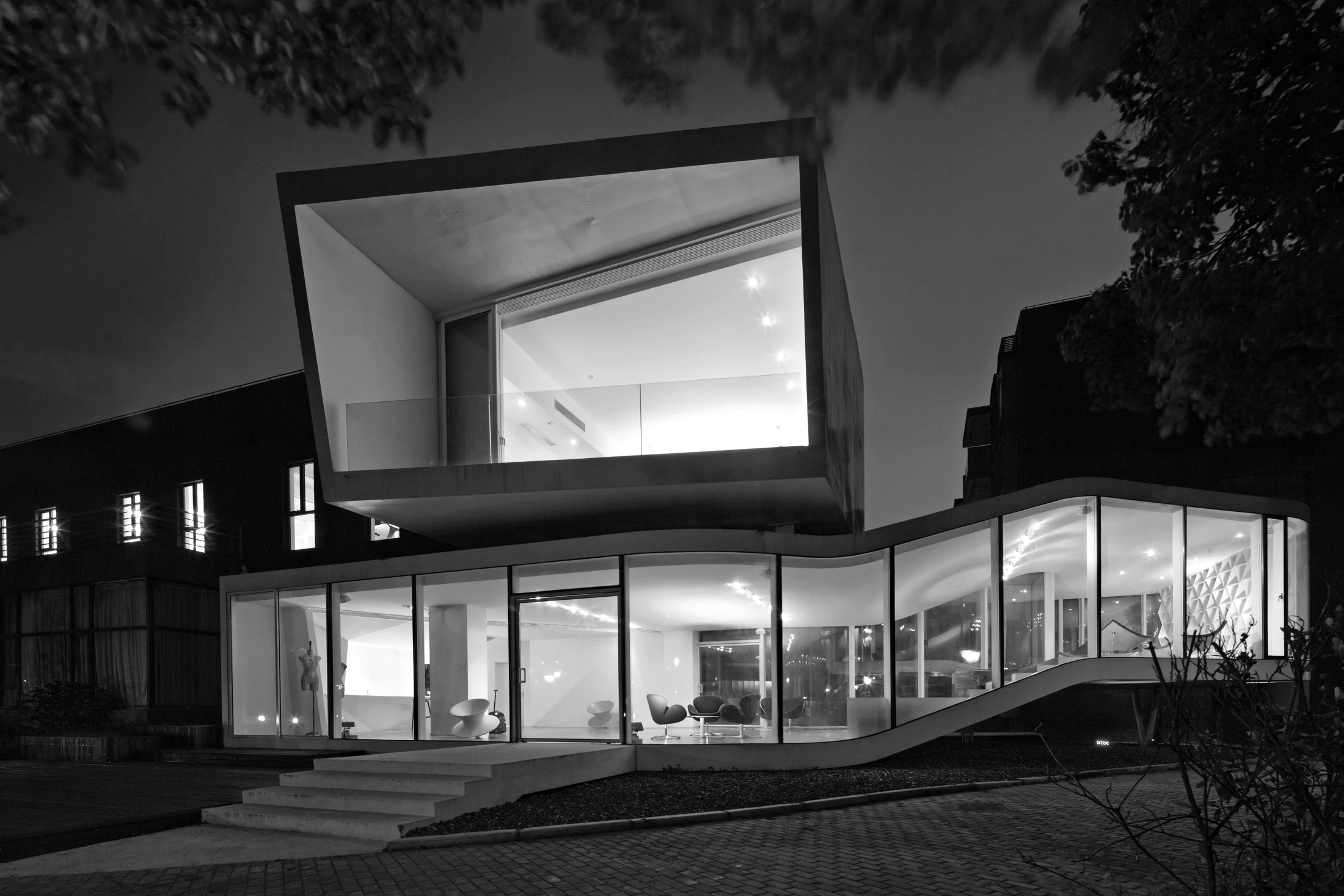 At night, the glazed frontage reveals the interior levels beneath the projecting upper room.