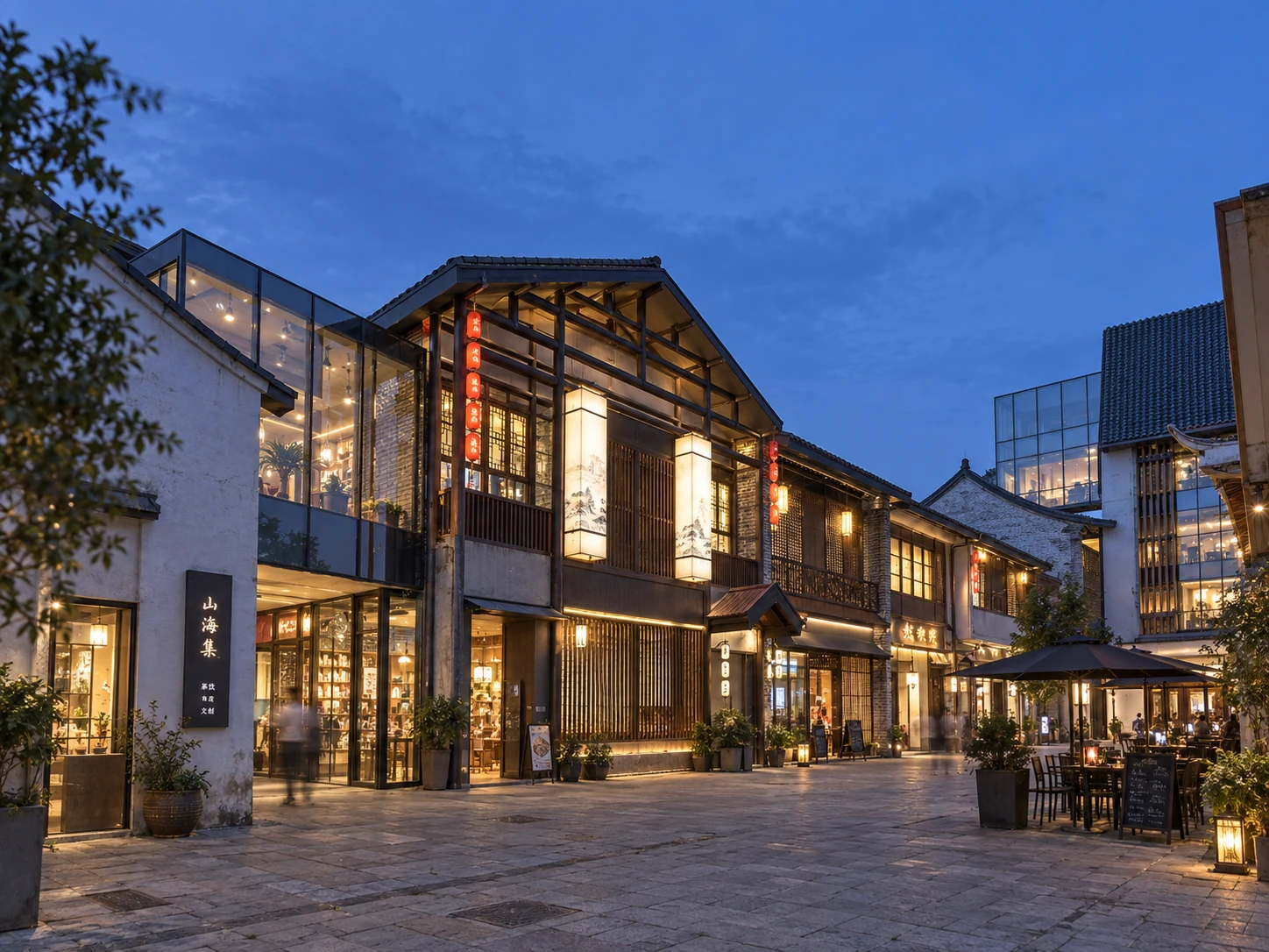 Completed courtyard at dusk with illuminated storefronts, pitched roofs, outdoor seating, and layered façades